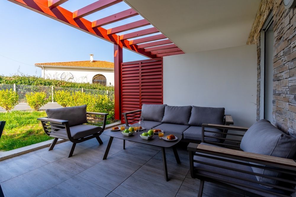 Outdoor patio with gray furniture, red trellis, fruit, and a building in the background.