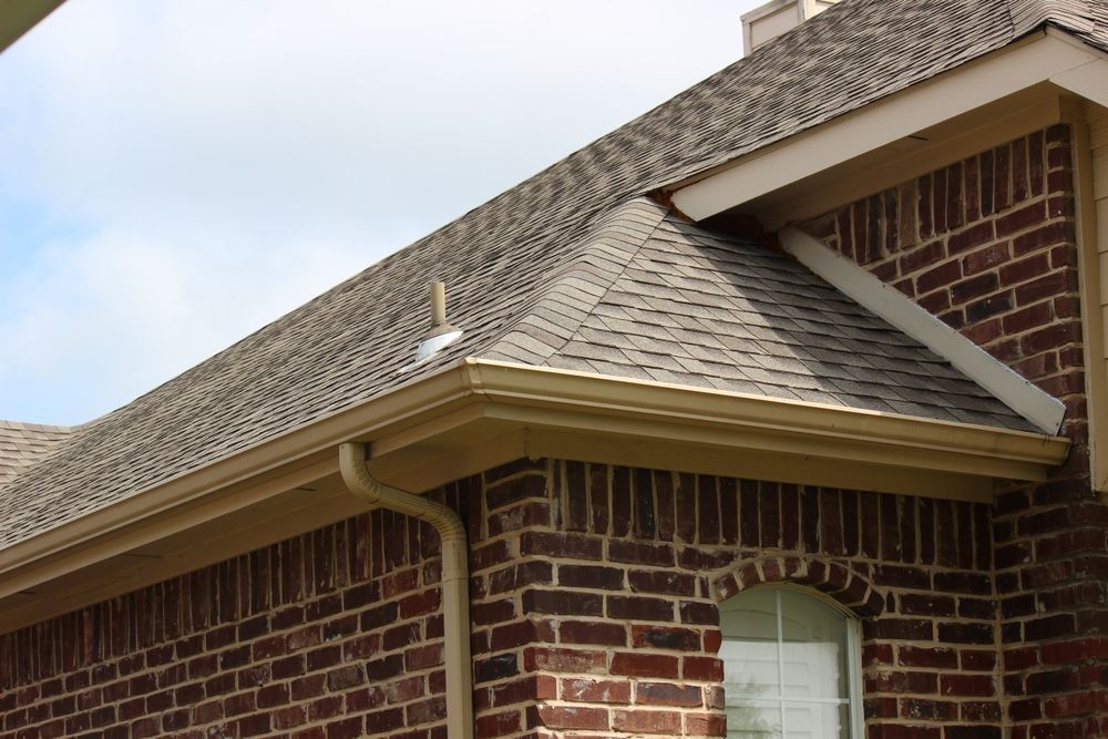 Brown brick house exterior with a shingled roof, beige gutters, and a cloudy sky.