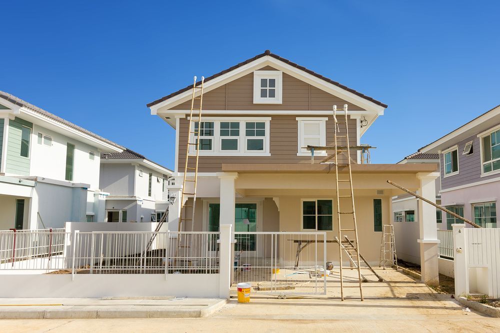 Two-story house under construction, tan siding, beige porch, blue sky. Ladders present.