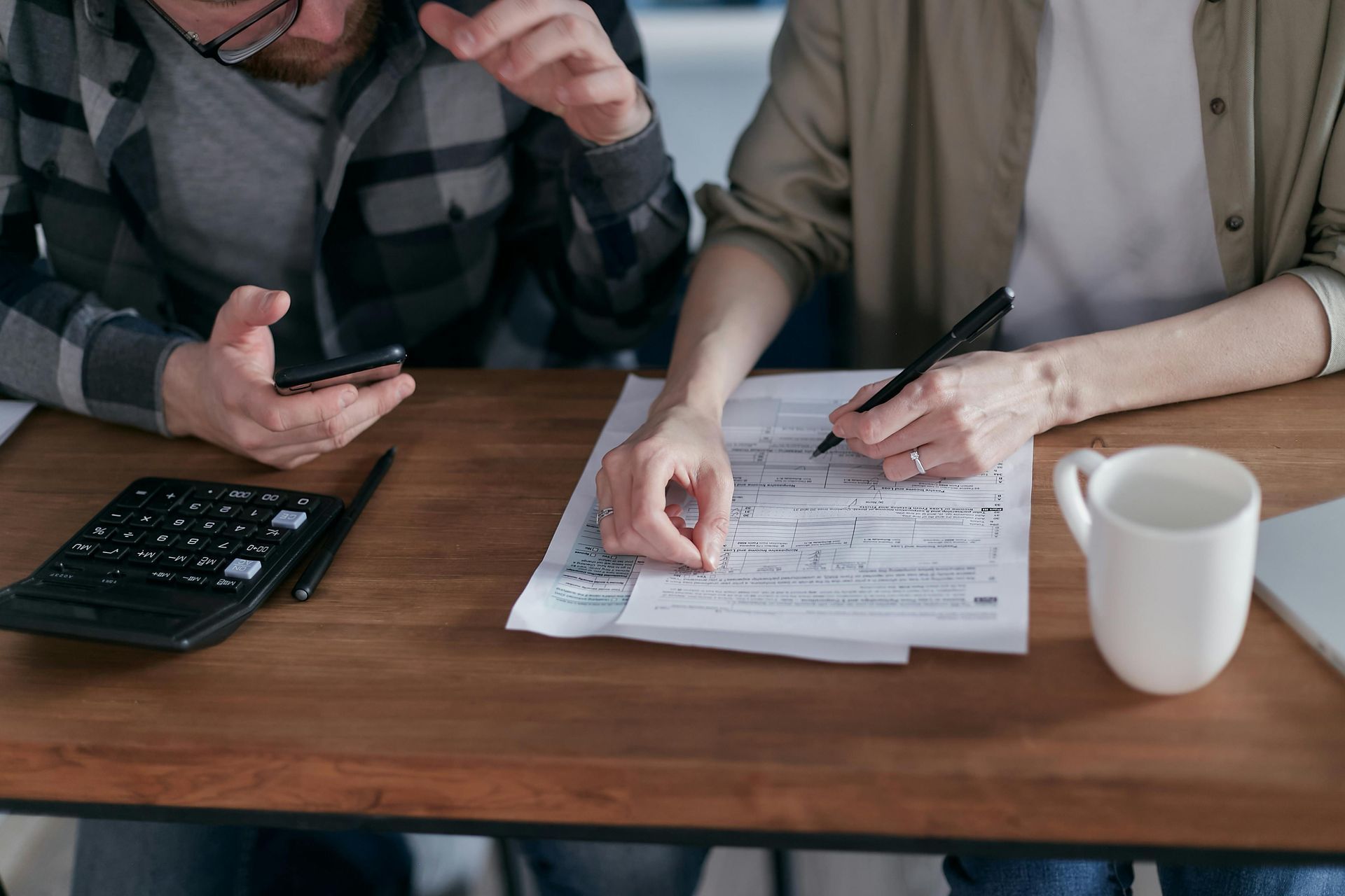 Two people working together at a wooden desk with a calculator, documents, a pen, and a white mug.