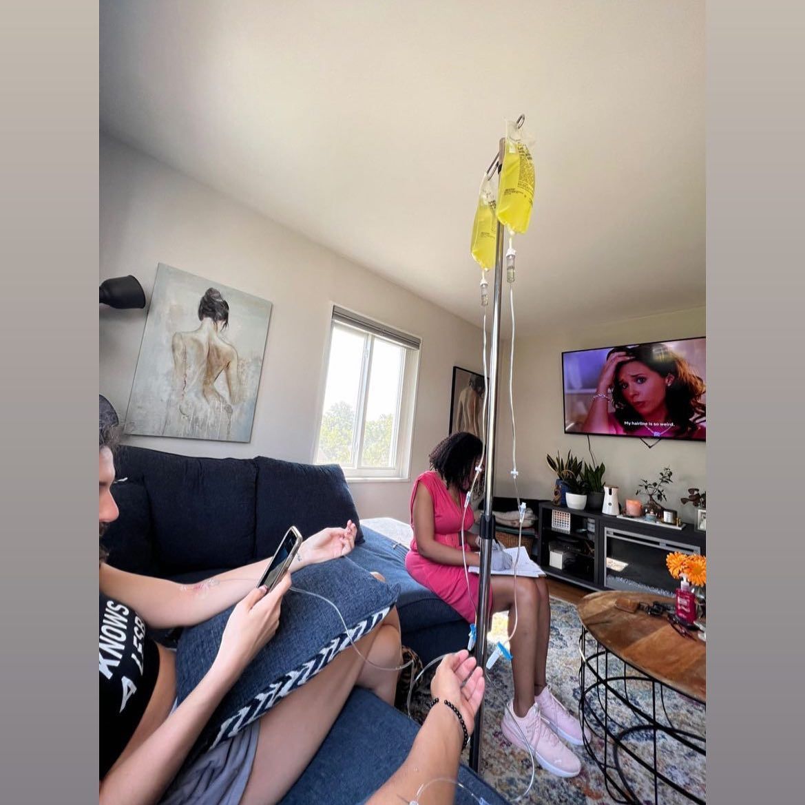 Two people in a living room, one receiving an IV drip. One on couch with phone, other watching TV.