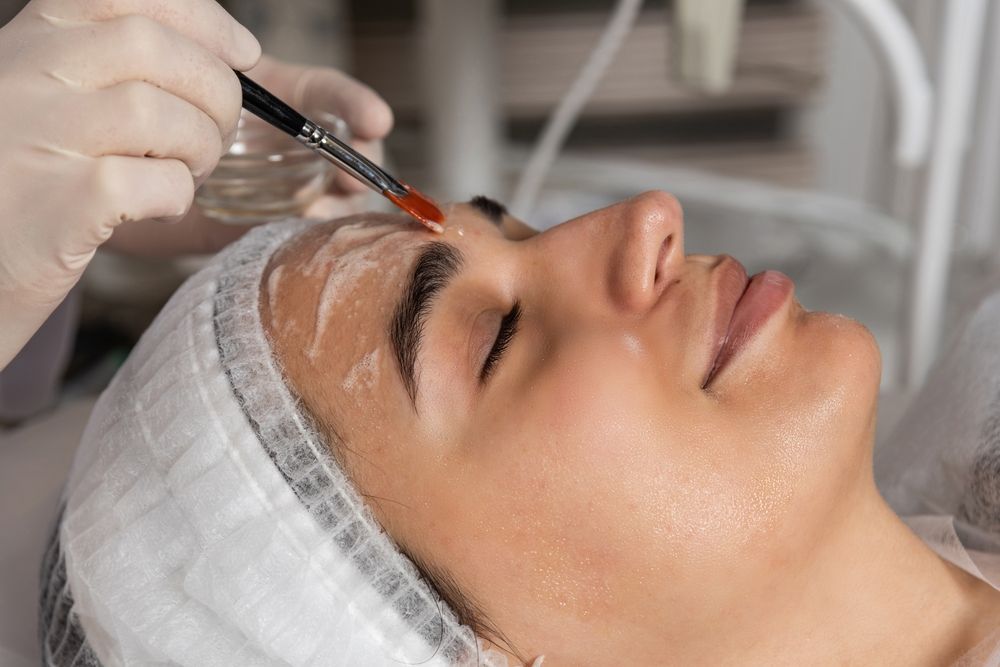 Woman receiving a facial peel. Technician applies product with brush. She's wearing a hair net and gloves.