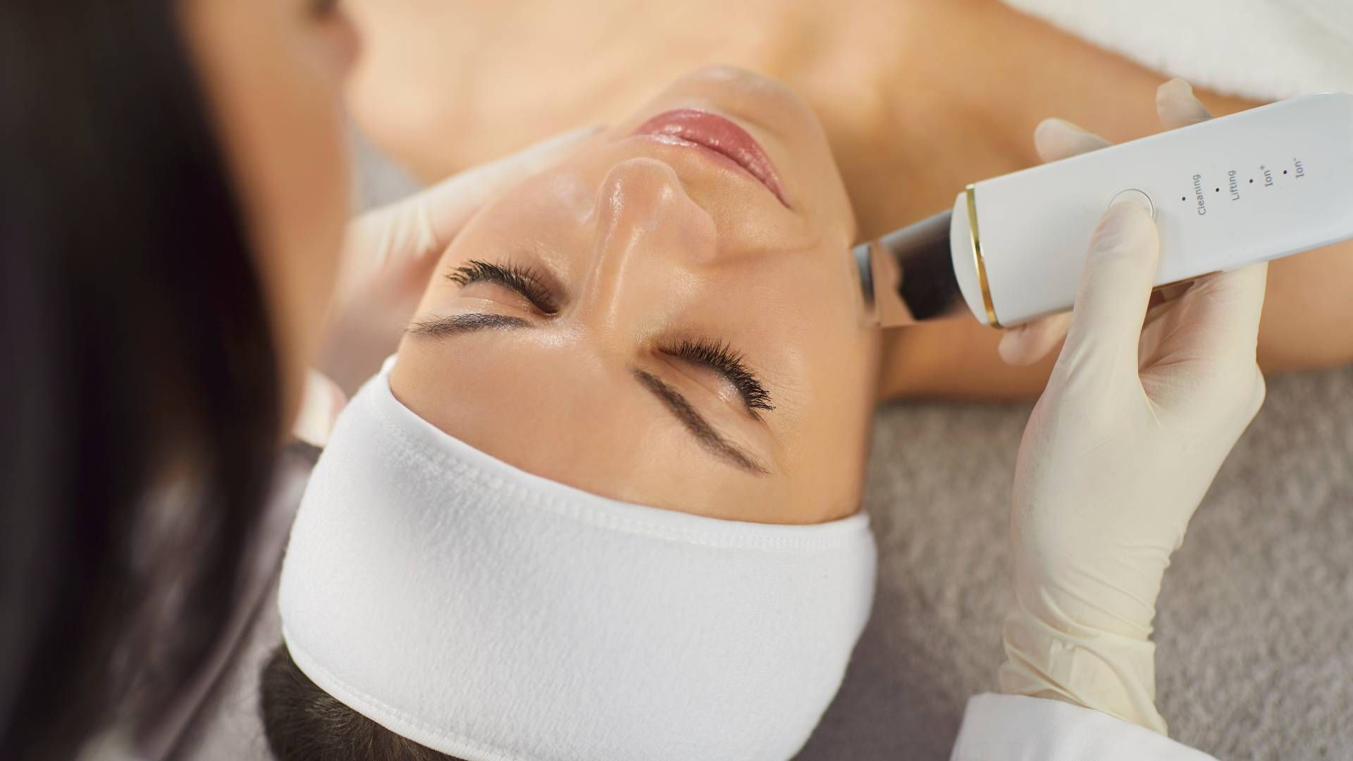 Person receiving facial treatment with a white device, lying on a table.