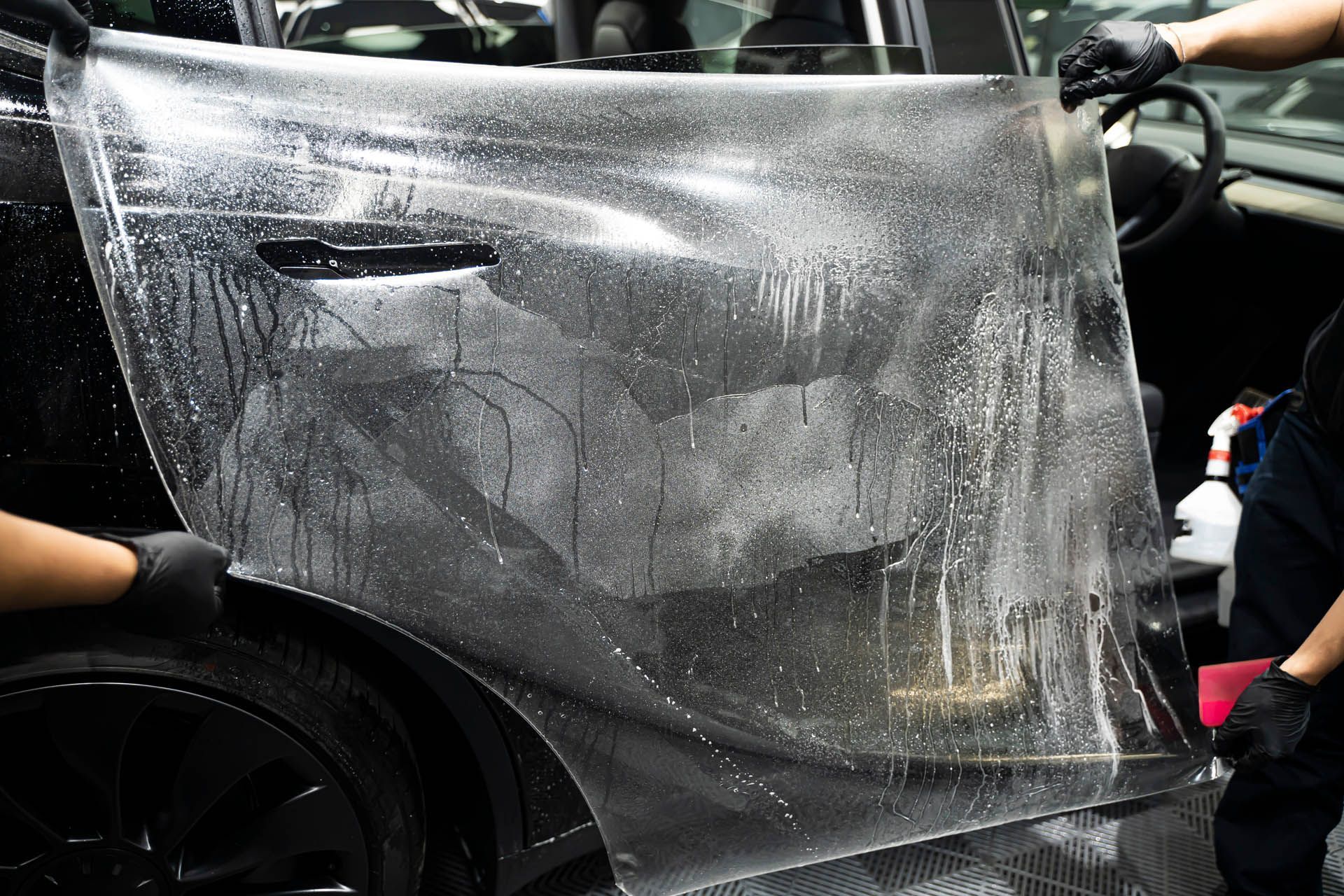 Applying clear protective film to a car door, wet with liquid, by two gloved hands.