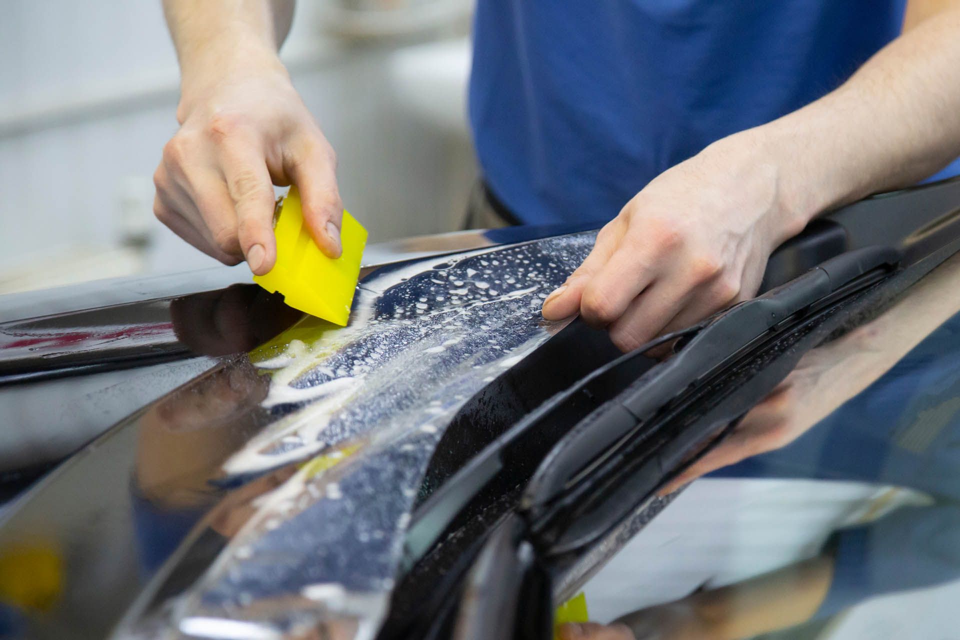 Hands smoothing window tint with a yellow squeegee on a car surface, reflecting the interior.