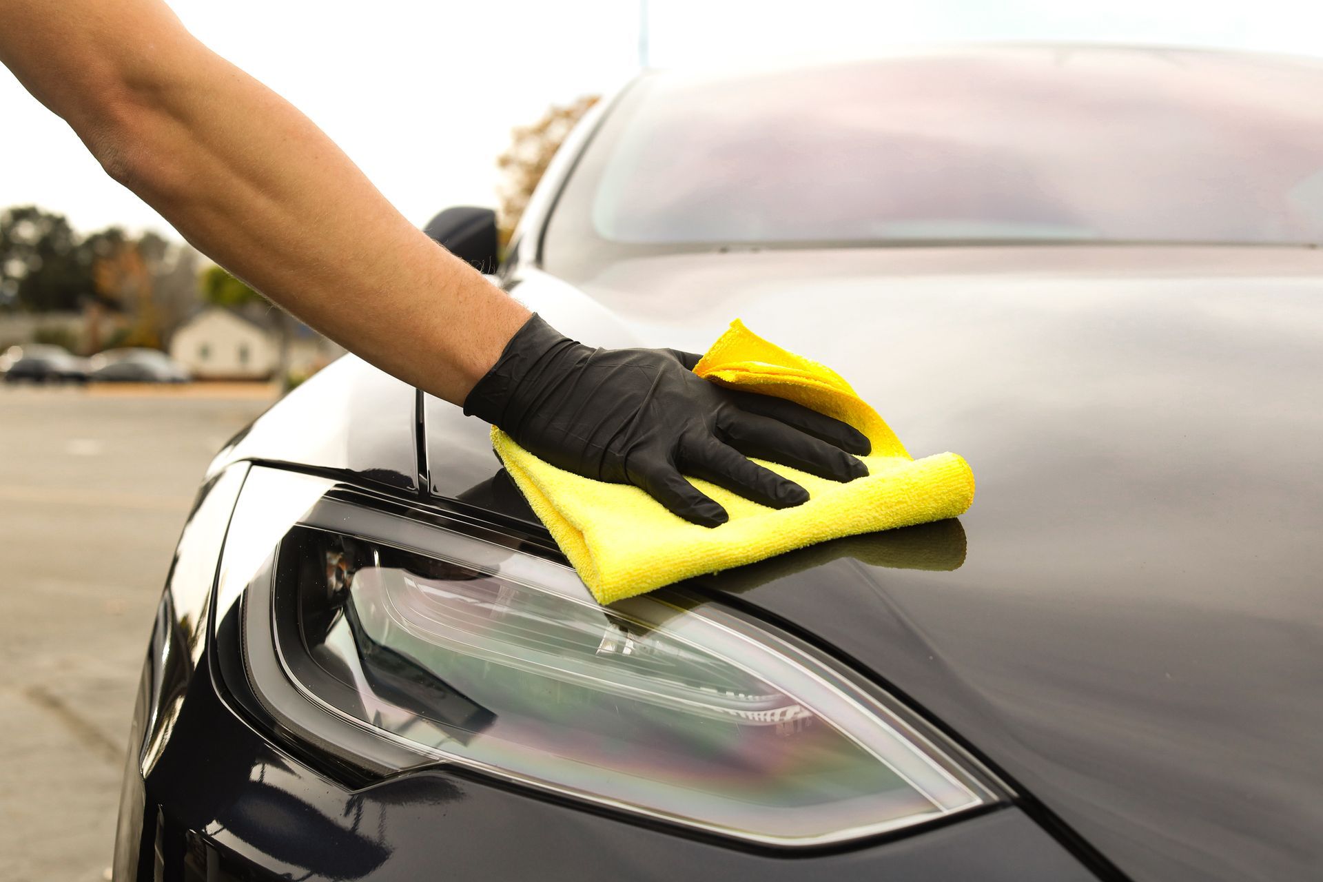 Person in black glove wiping a black car's hood with a yellow cloth outdoors.