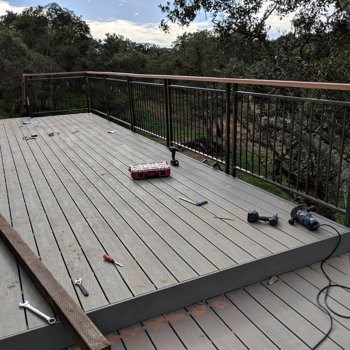 Deck with gray composite decking, black railing, and tools, surrounded by trees.