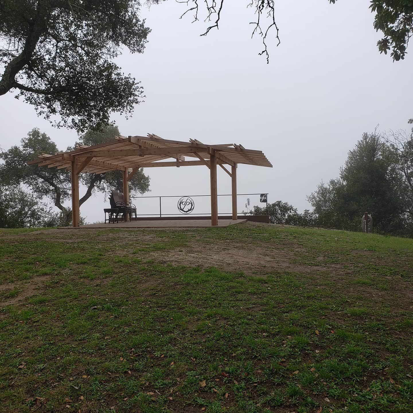 A person sits on a bench under a wooden gazebo on a grassy hill, with a foggy backdrop.