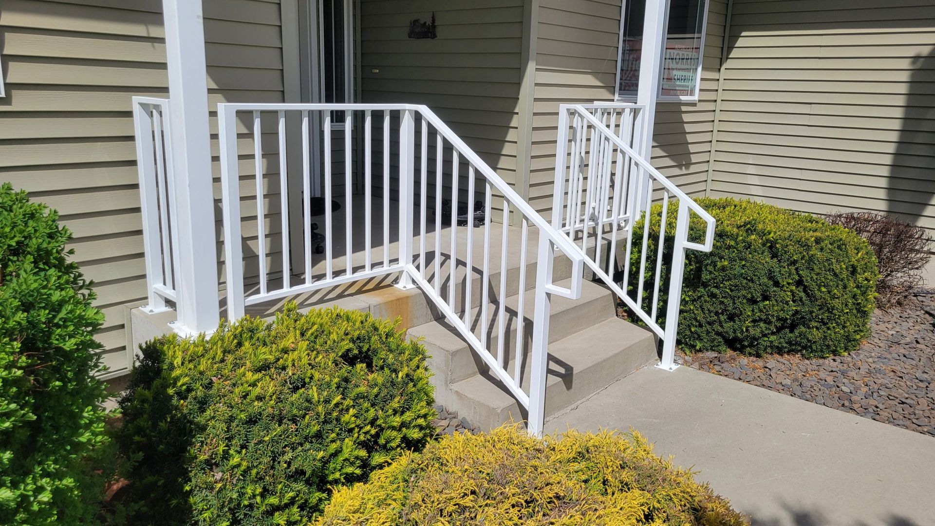 White railing surrounds concrete steps leading to a front door, flanked by green bushes.
