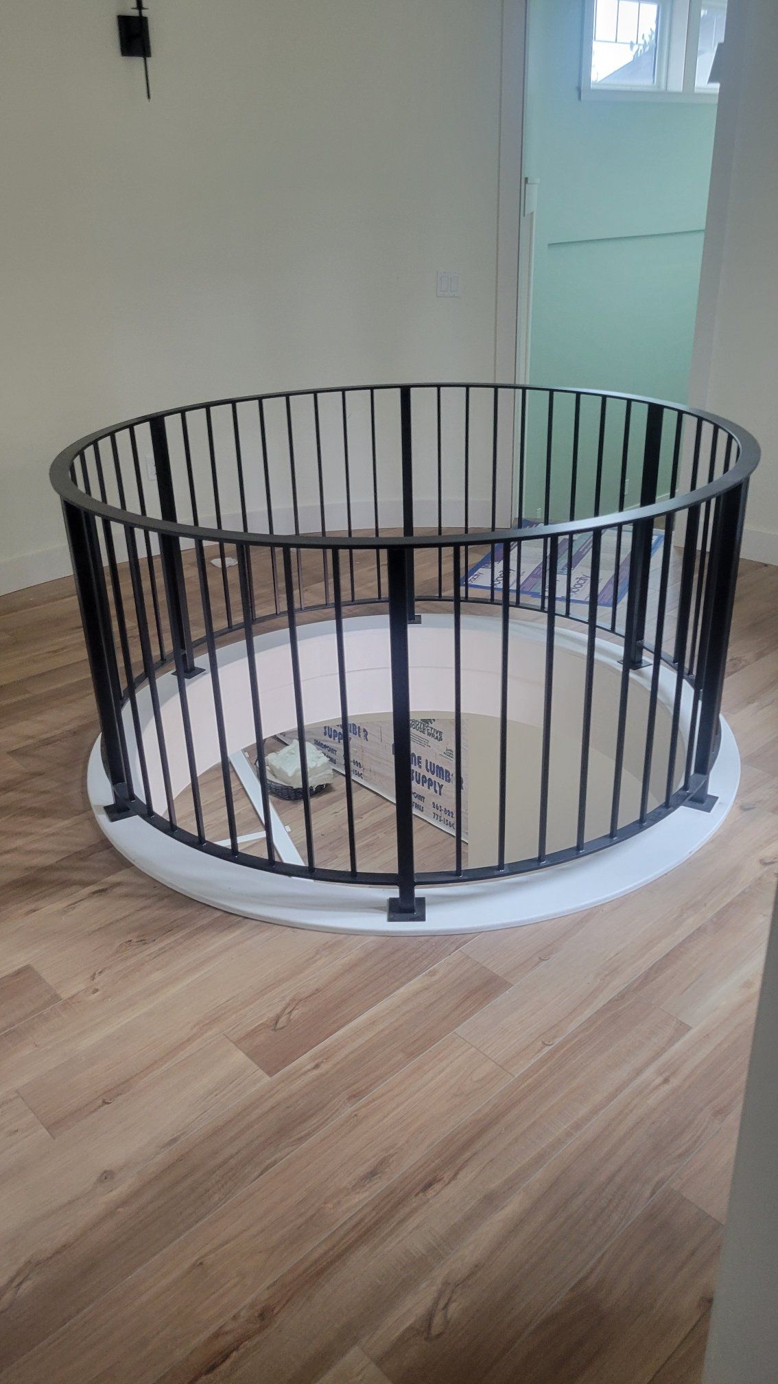 Black circular railing on a white platform, protecting a circular opening in the floor, in a room with light brown flooring.