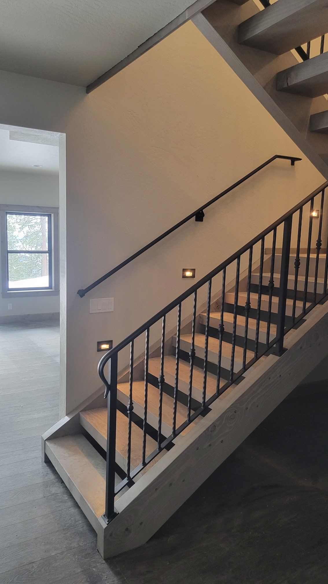 Staircase with black railings and recessed lighting. Cream walls, light wood stairs, and a window in the background.