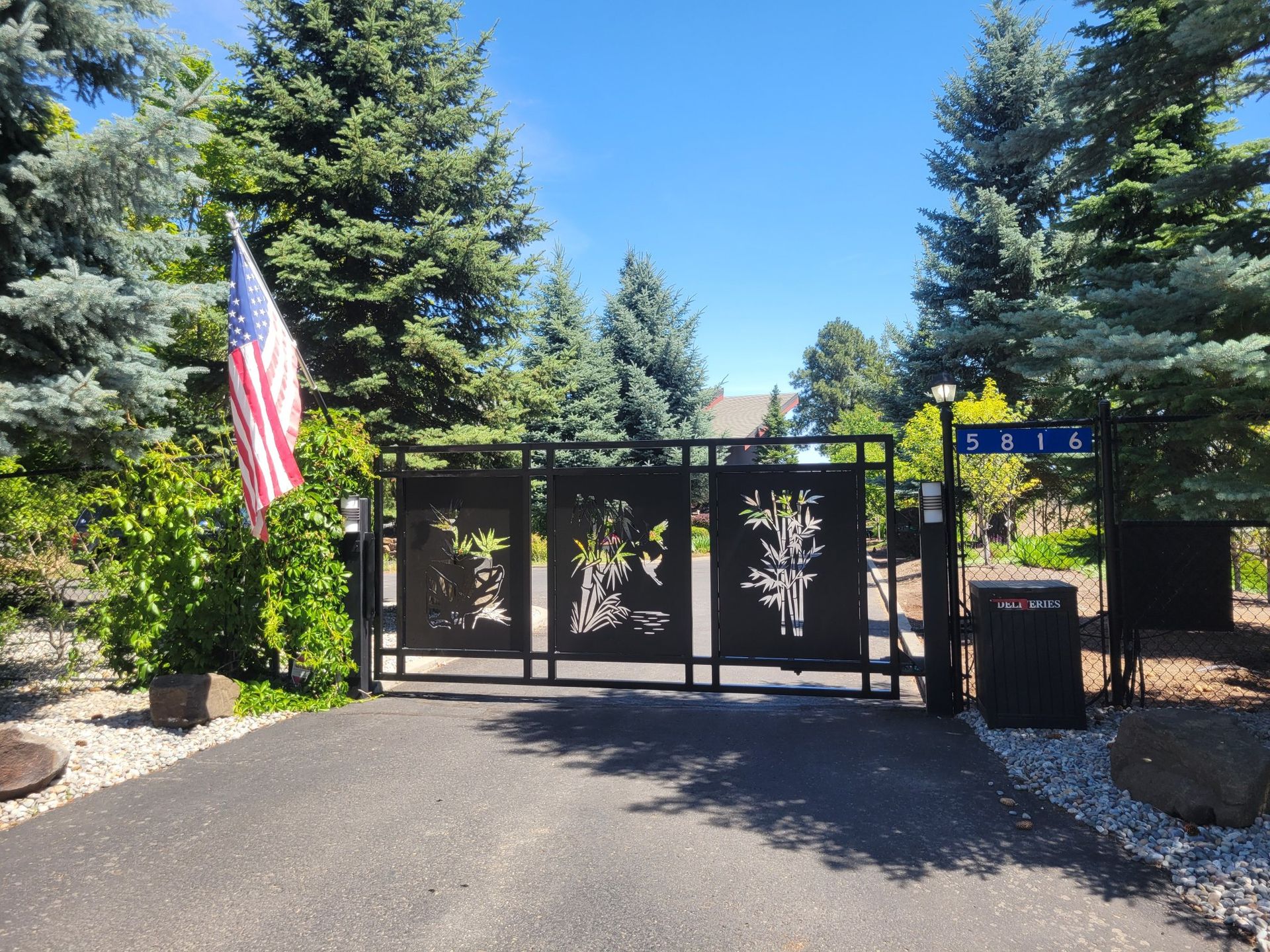 Black metal gate with tree designs, driveway, American flag, house number sign, and lush greenery under a blue sky.
