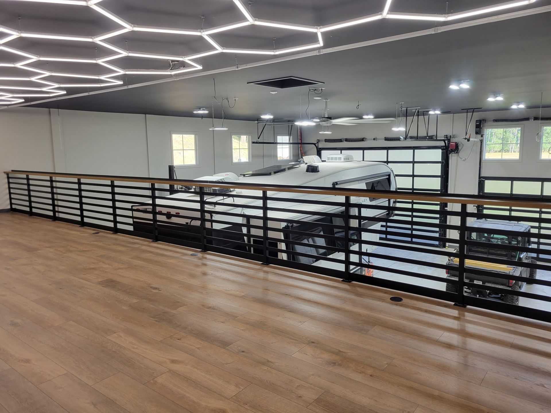 Wooden balcony with black railing overlooking a garage with a white car. Hexagon ceiling lights.