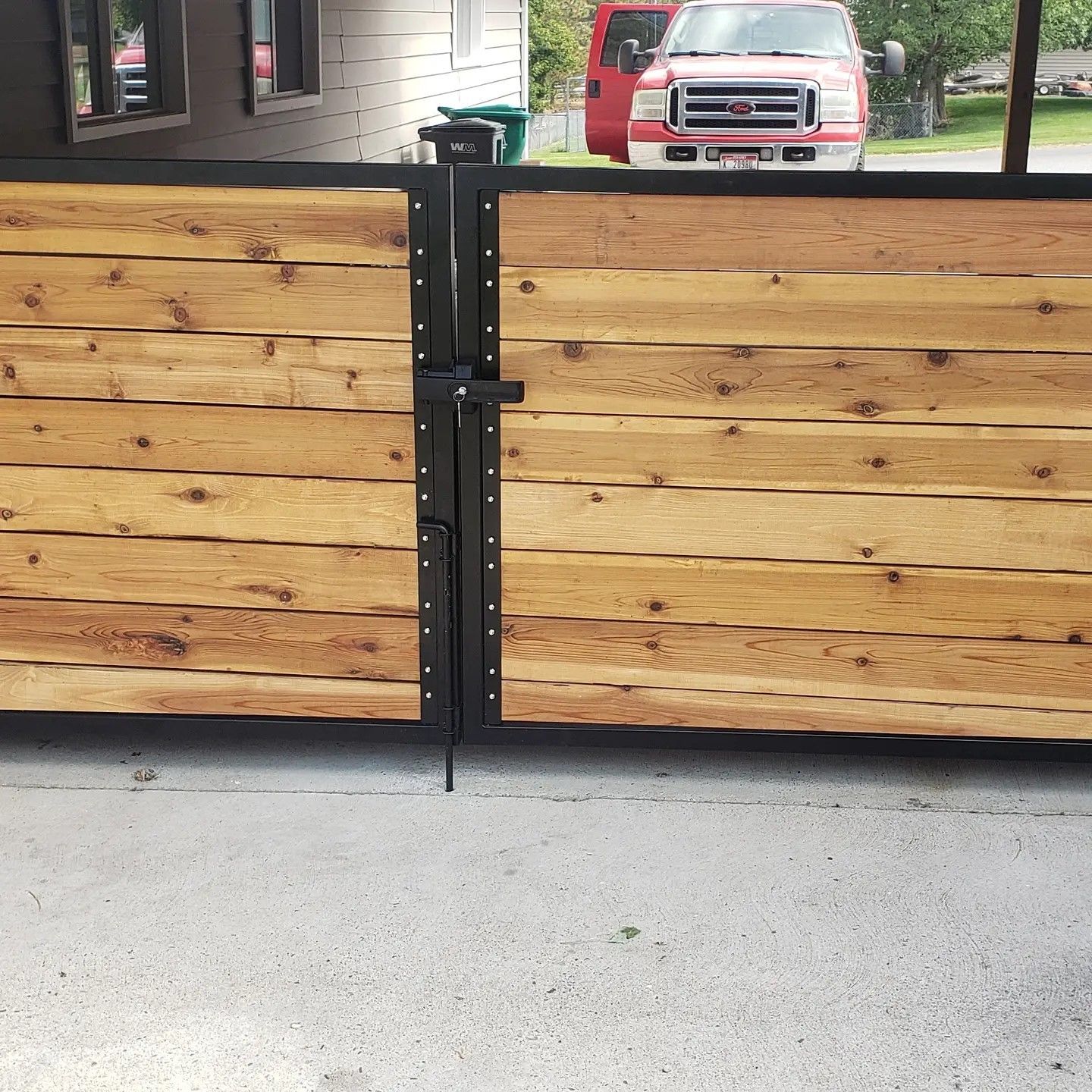 Wooden gate with black metal frame and latch, set on concrete, with a red truck visible.