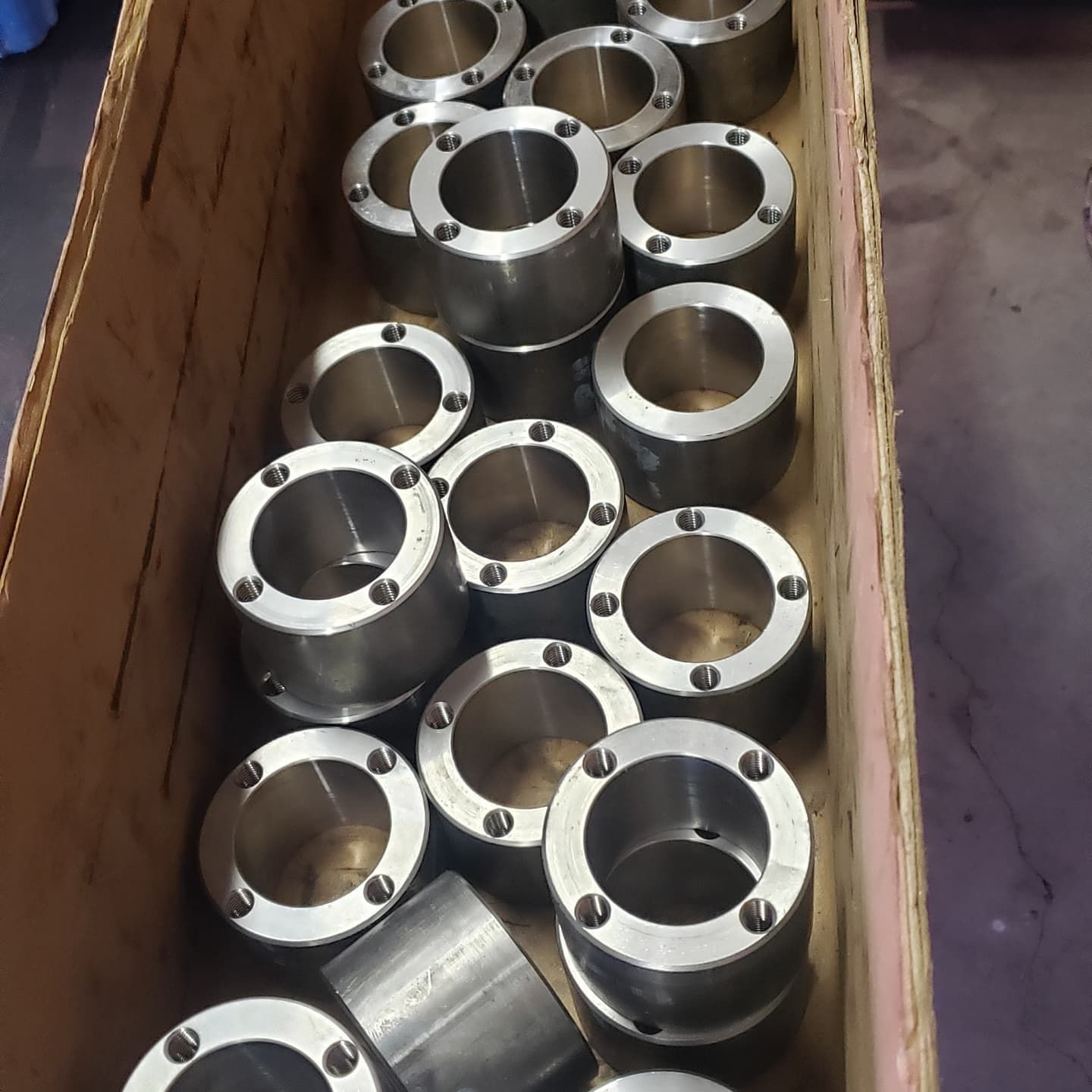 A wooden crate filled with metal cylindrical parts with circular holes around the edges.