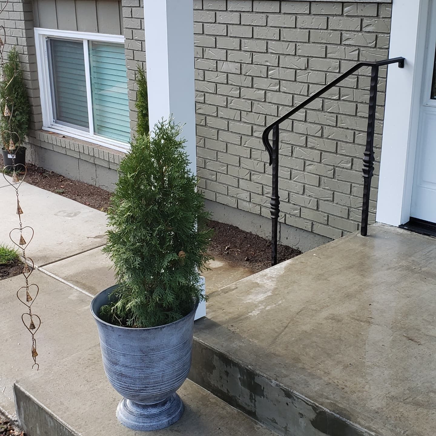 A potted evergreen tree sits on a concrete porch next to a brick wall and a white pillar.