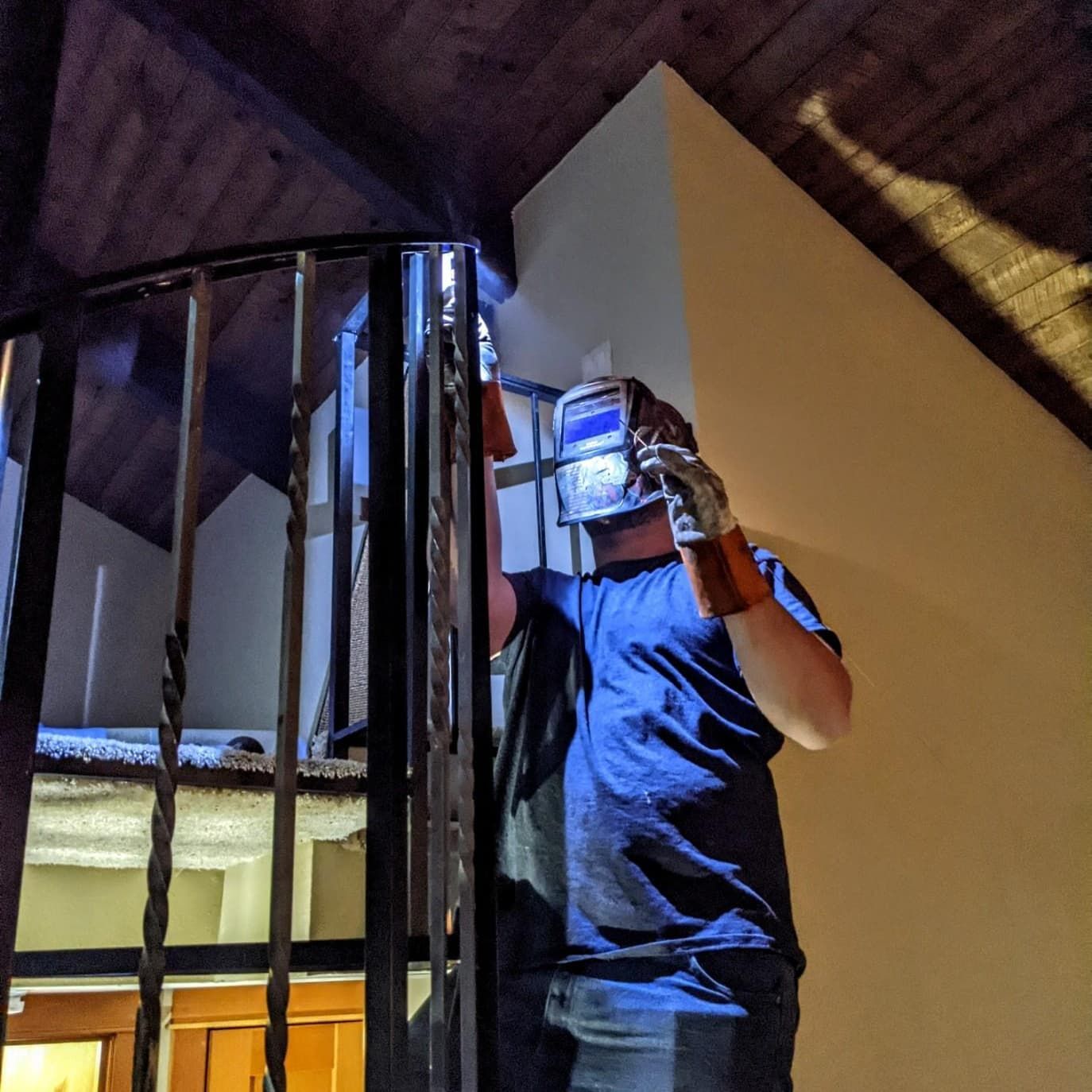 Person welding metal handrail, wearing a welding mask and gloves, indoors, near a stairwell.