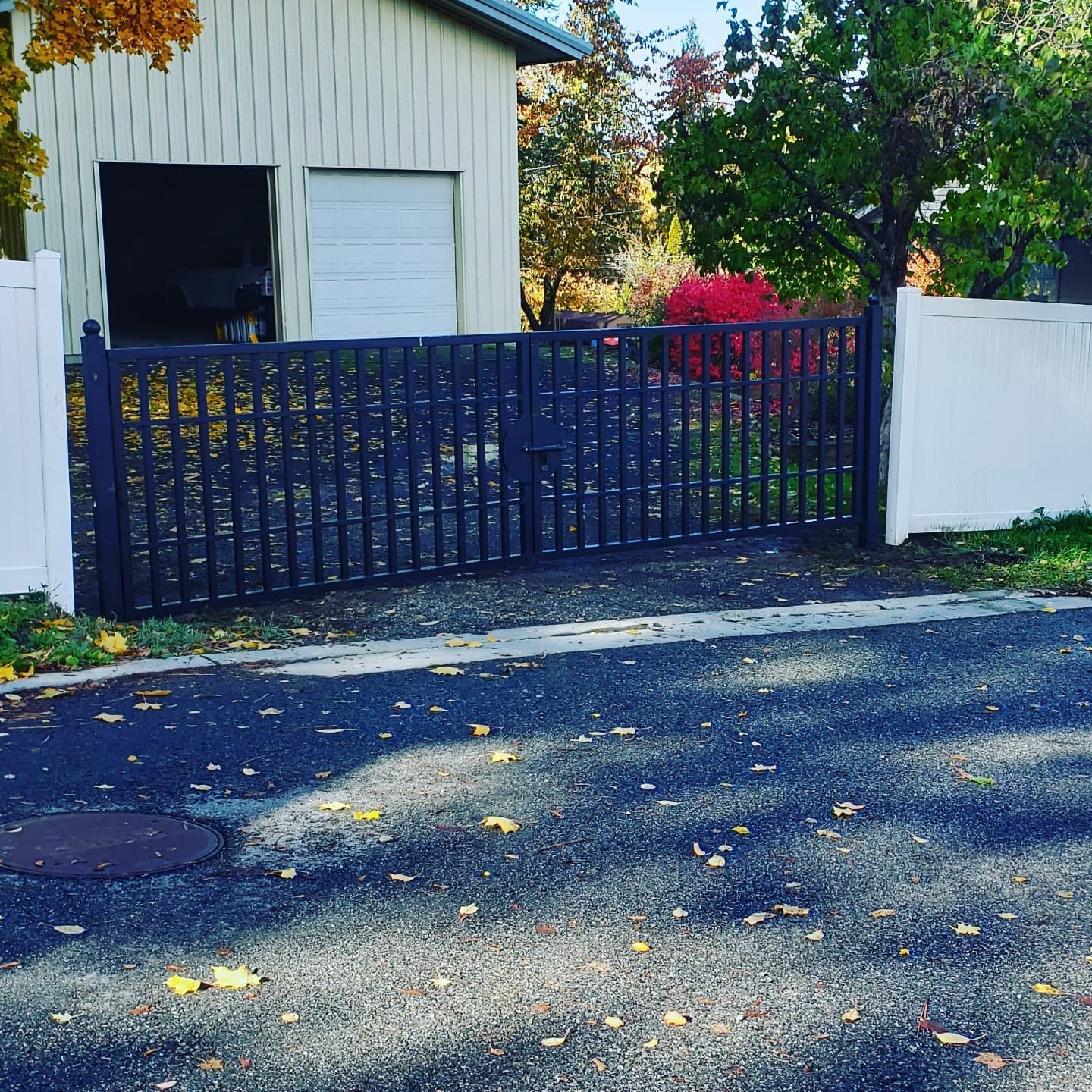 Black gate with vertical bars between white fences, garage in the background, autumn leaves on pavement.