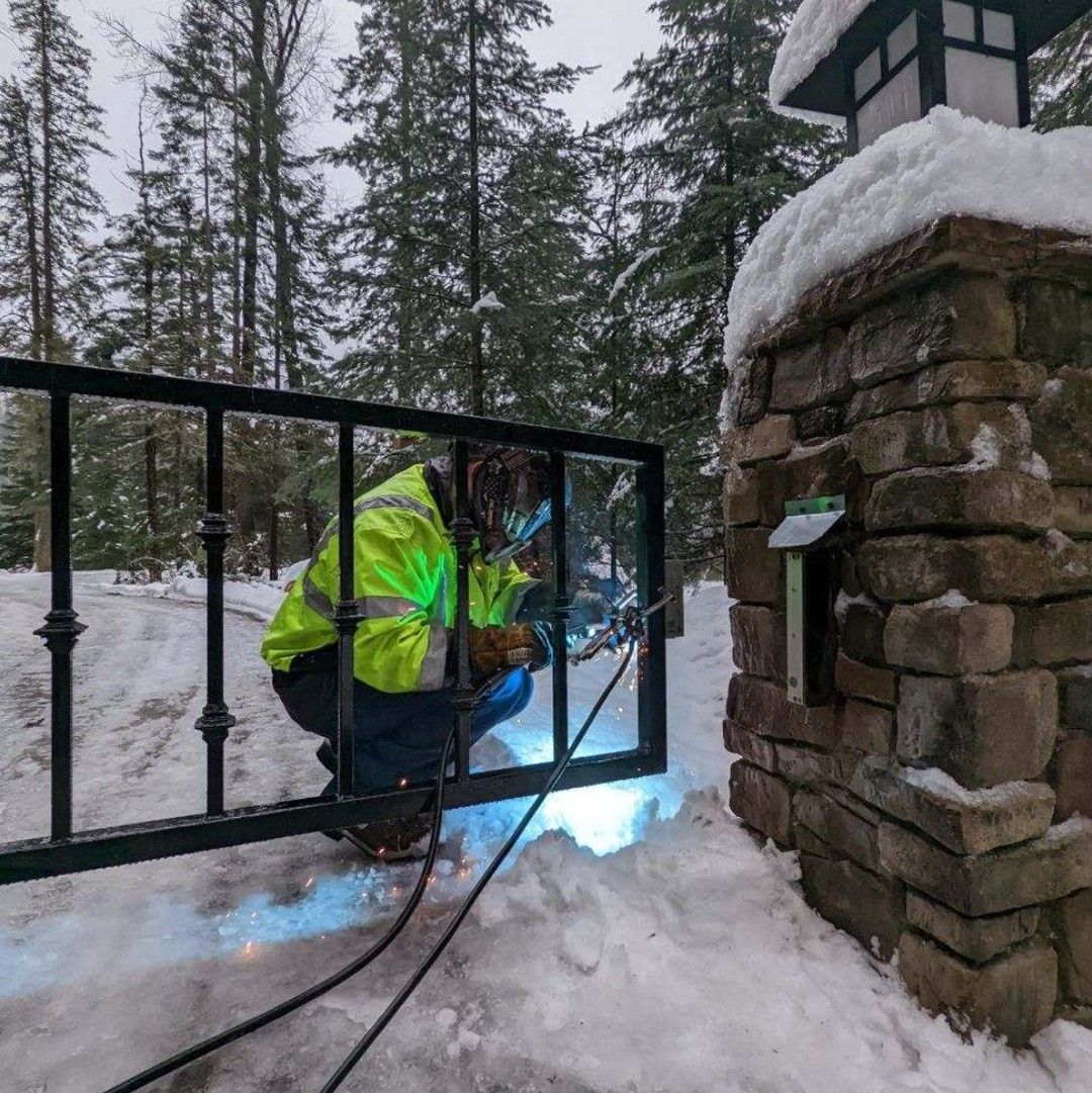 Welder in a high-visibility jacket welding a black gate in a snowy outdoor setting.