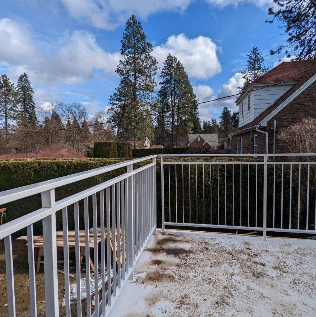 White railing on a weathered balcony overlooking a hedge, trees, and house with a red roof under a cloudy sky.