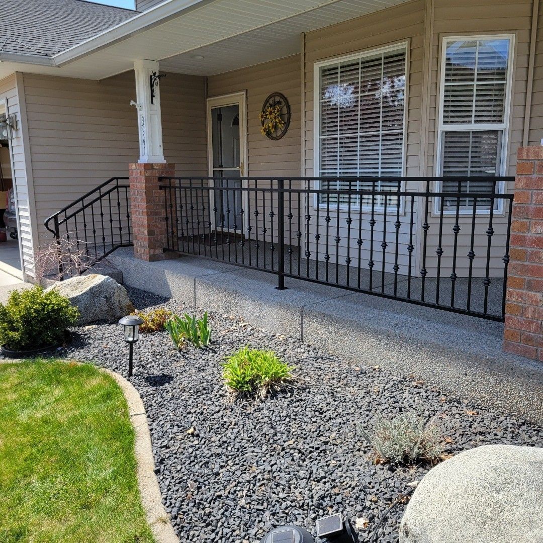 Exterior view of a house with a porch. Black iron railing, brick columns, and a stone landscape with plants.