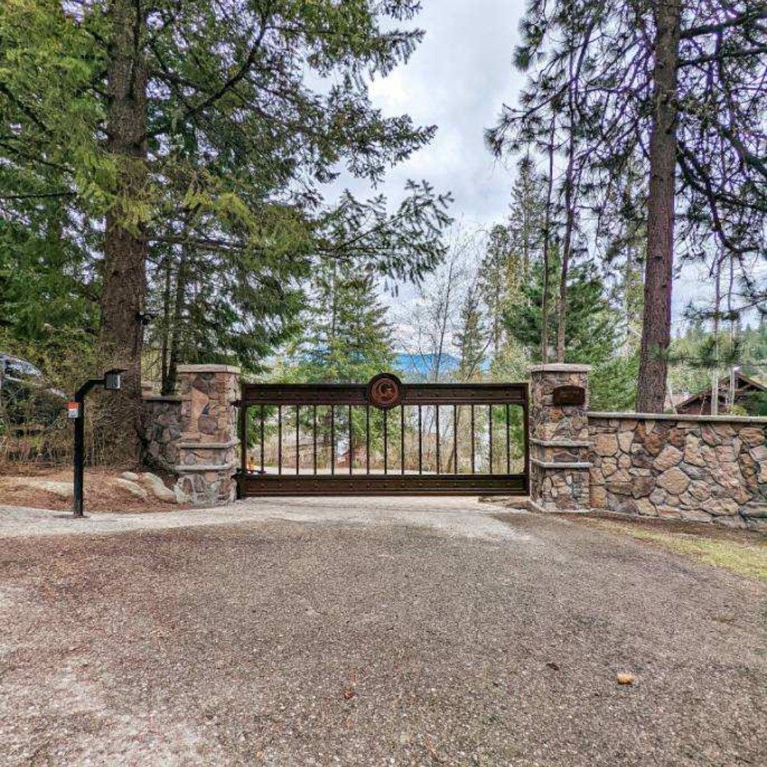 Gated driveway entrance with stone pillars, gravel, trees, and partial view of a lake.