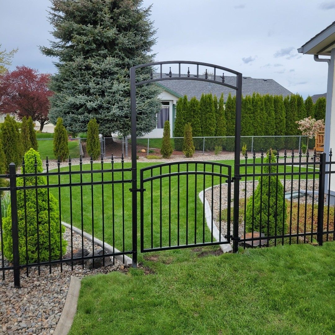 Black wrought-iron fence with gate and archway in front yard, with green grass and trees in background.