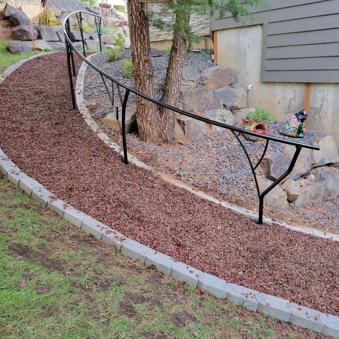 A curved pathway with black railing through a yard, bordered by stone and filled with wood chips.