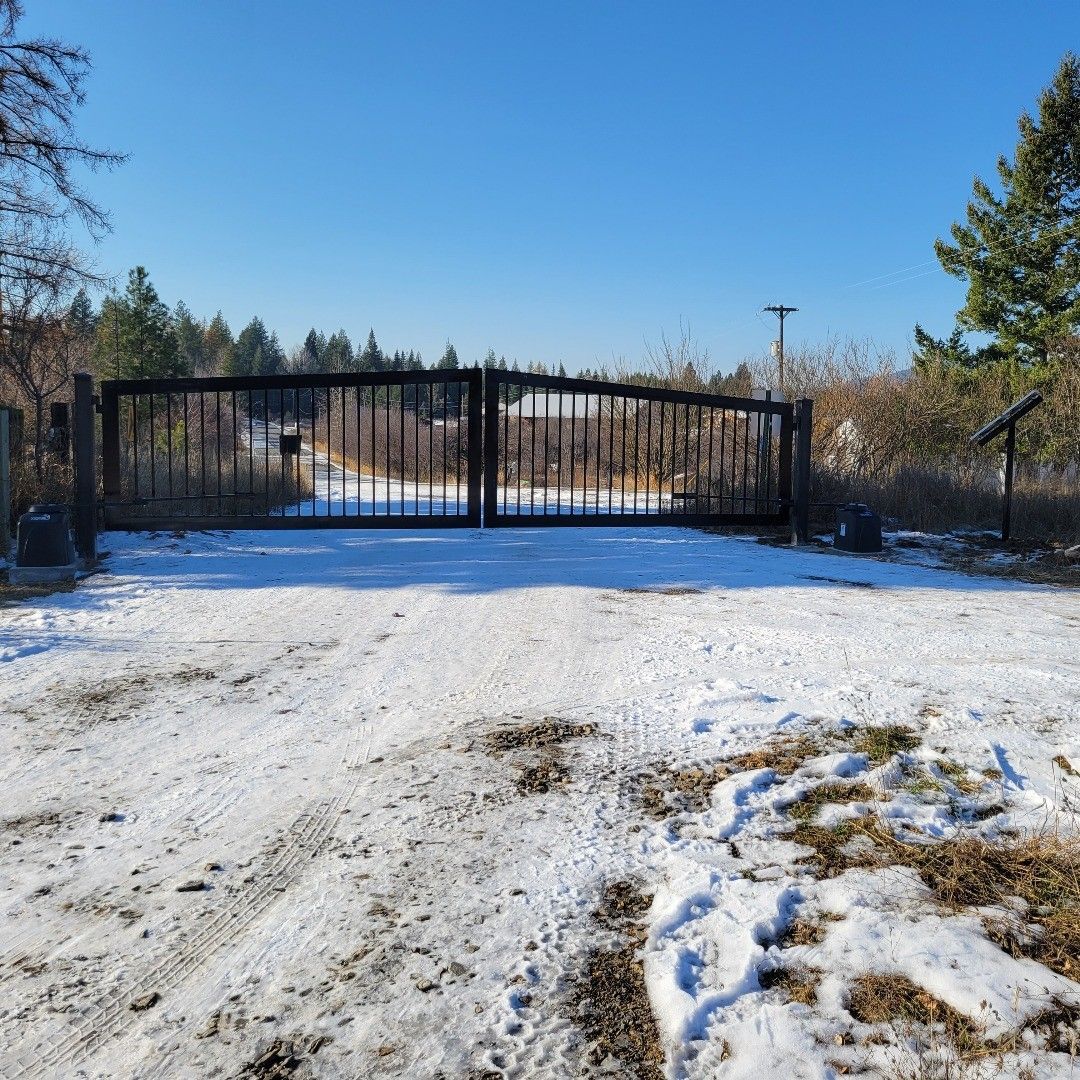 Snowy driveway leads to black gate and distant trees under a bright blue sky.