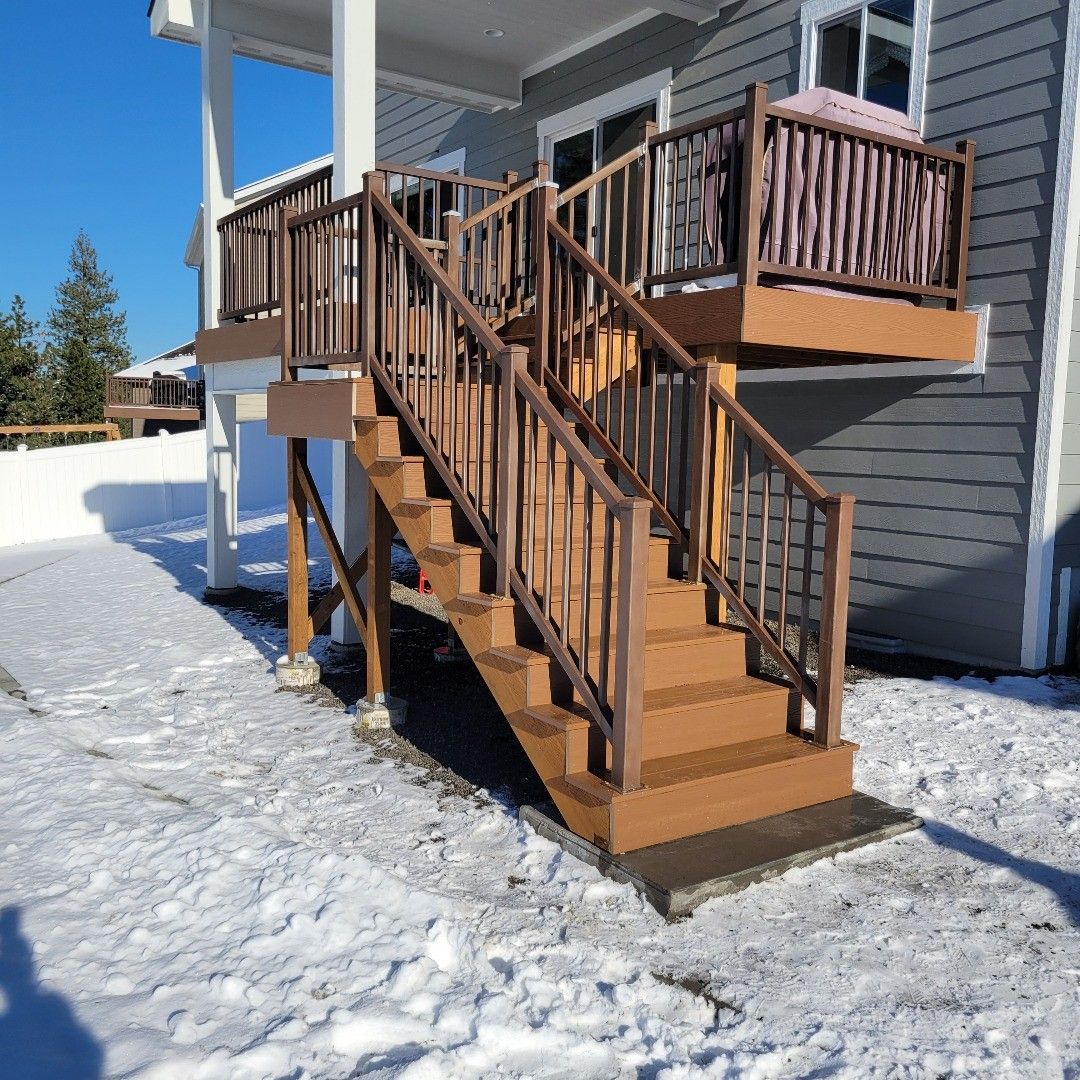 Brown wooden deck with stairs in a snowy yard, next to a gray house.