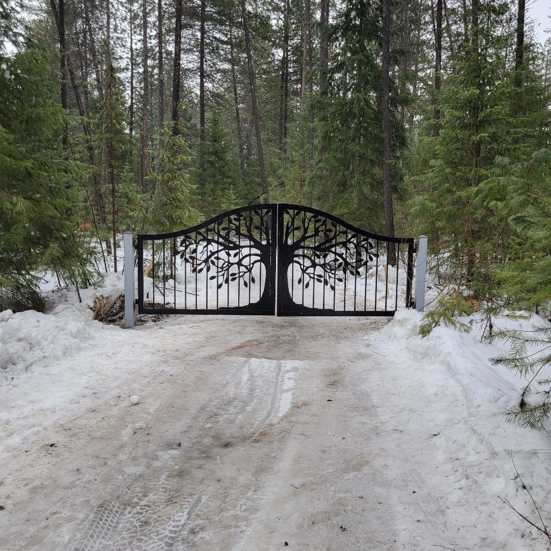 Black metal gate with tree design, set in snowy driveway, forest background.