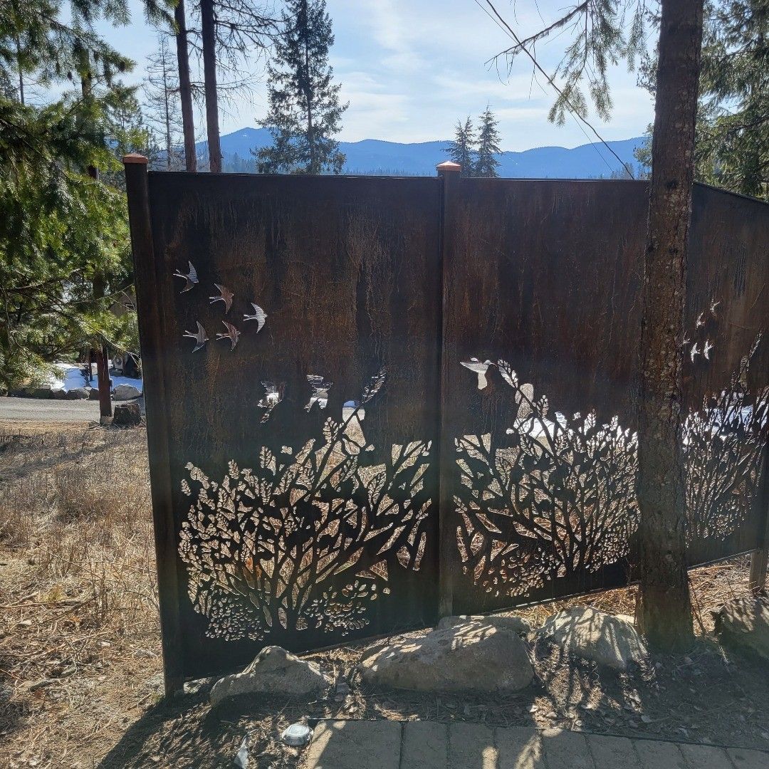 Metal art gate with cut-out bird and tree silhouettes, against a background of trees and mountains.