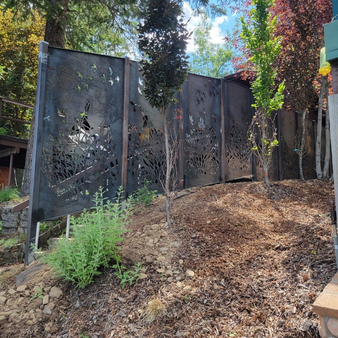 Black metal decorative fence with tree-shaped cutouts on a hillside with trees and plants.