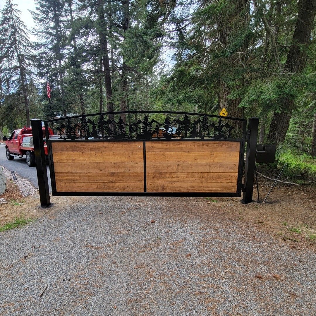Wooden gate with black metal detail, set at a forest entrance. A red truck is parked nearby.
