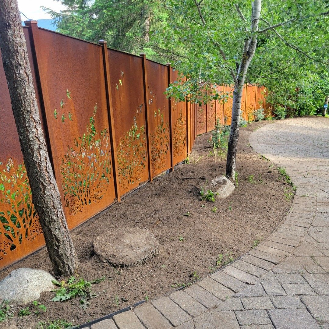 Rusty metal fence with floral cutouts alongside a brick path and trees.