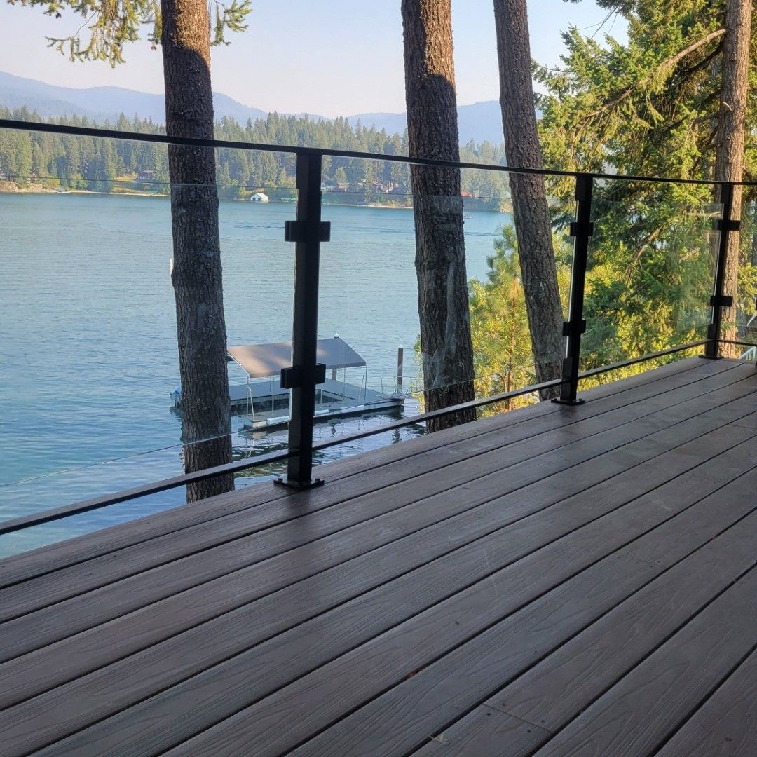 Wooden deck overlooking lake with glass and black railing, trees, and dock.