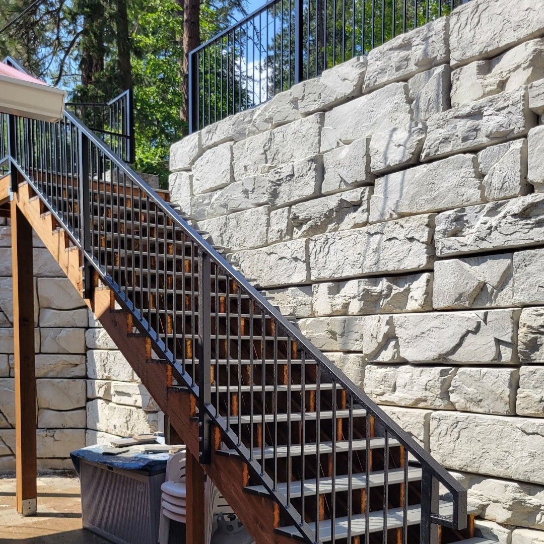 Wooden staircase with black railings against a large stone retaining wall.