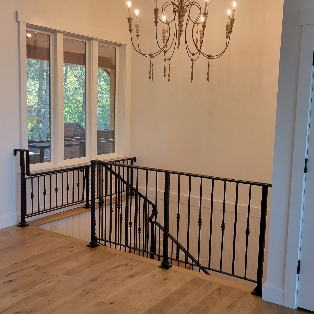 Stairwell with black metal railing, wooden floor, chandelier, and a large window overlooking a wooded area.