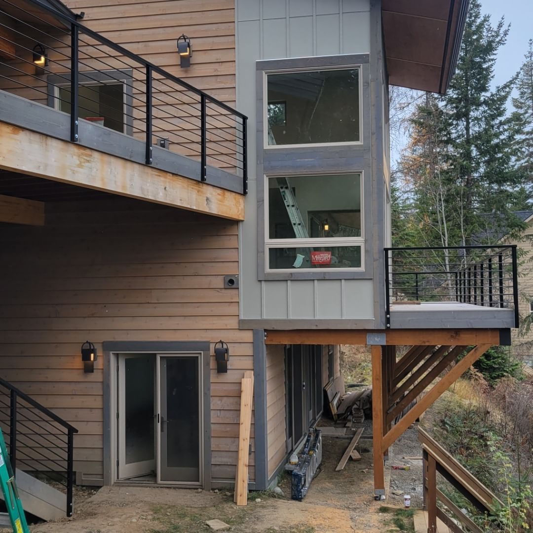 Side view of a house with decks, windows, and black railings. Brown siding with a wooden deck and supports.