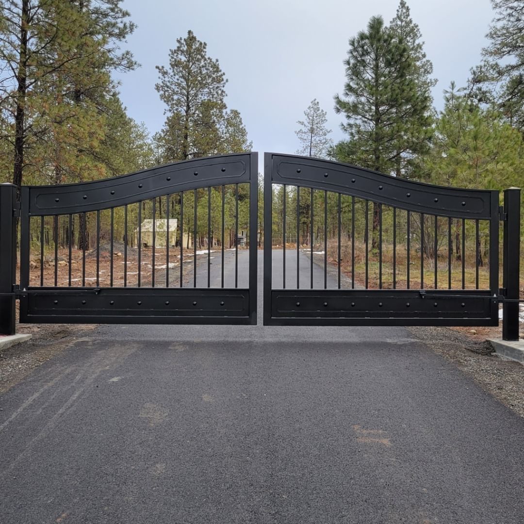 Black wrought-iron driveway gate, arched top, open on asphalt road, trees in the background.