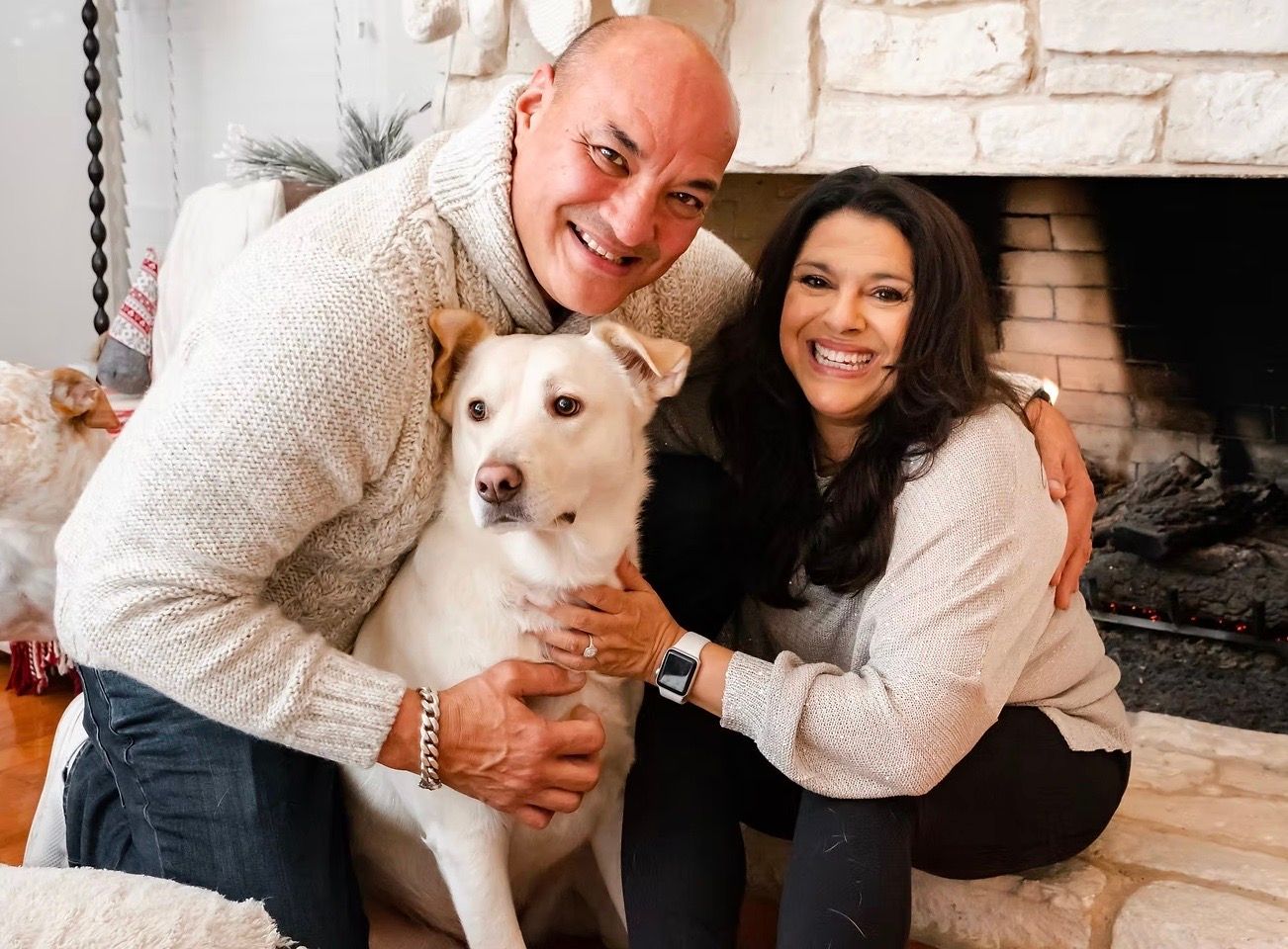 Man and woman embrace a yellow Labrador dog in front of a fireplace, all wearing sweaters.