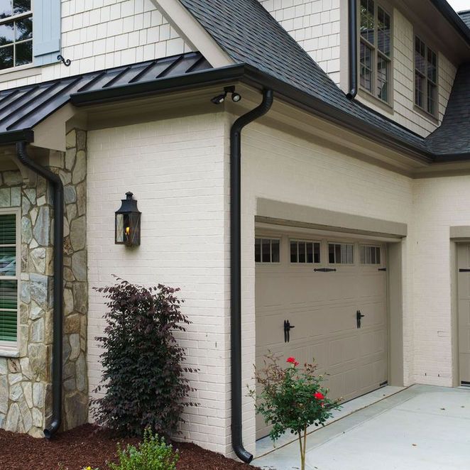 Tan-colored house exterior with black gutters, a garage door, a light fixture, and landscaping.