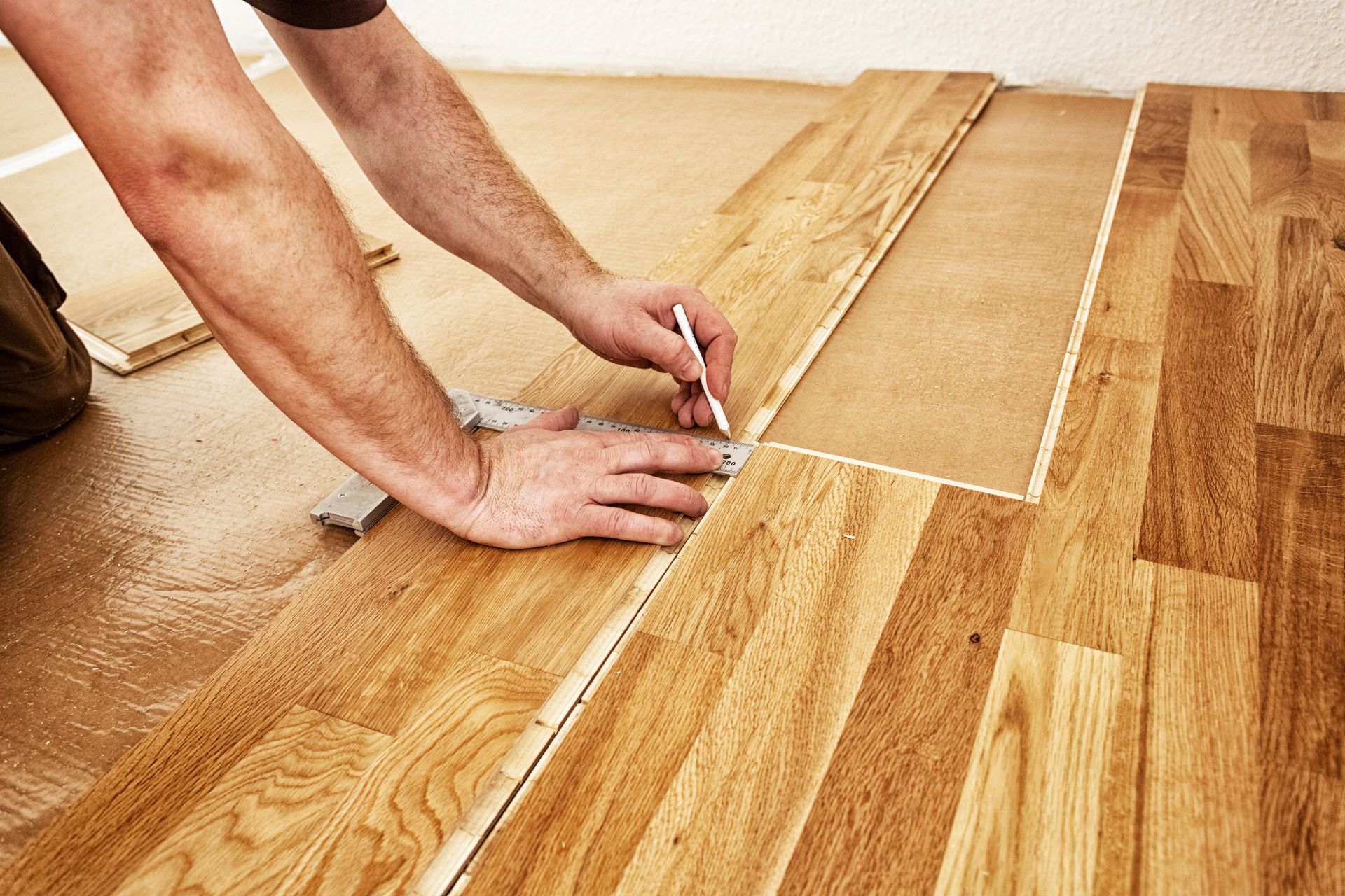 Person installing wooden flooring, using a pencil and square to measure and mark a plank.