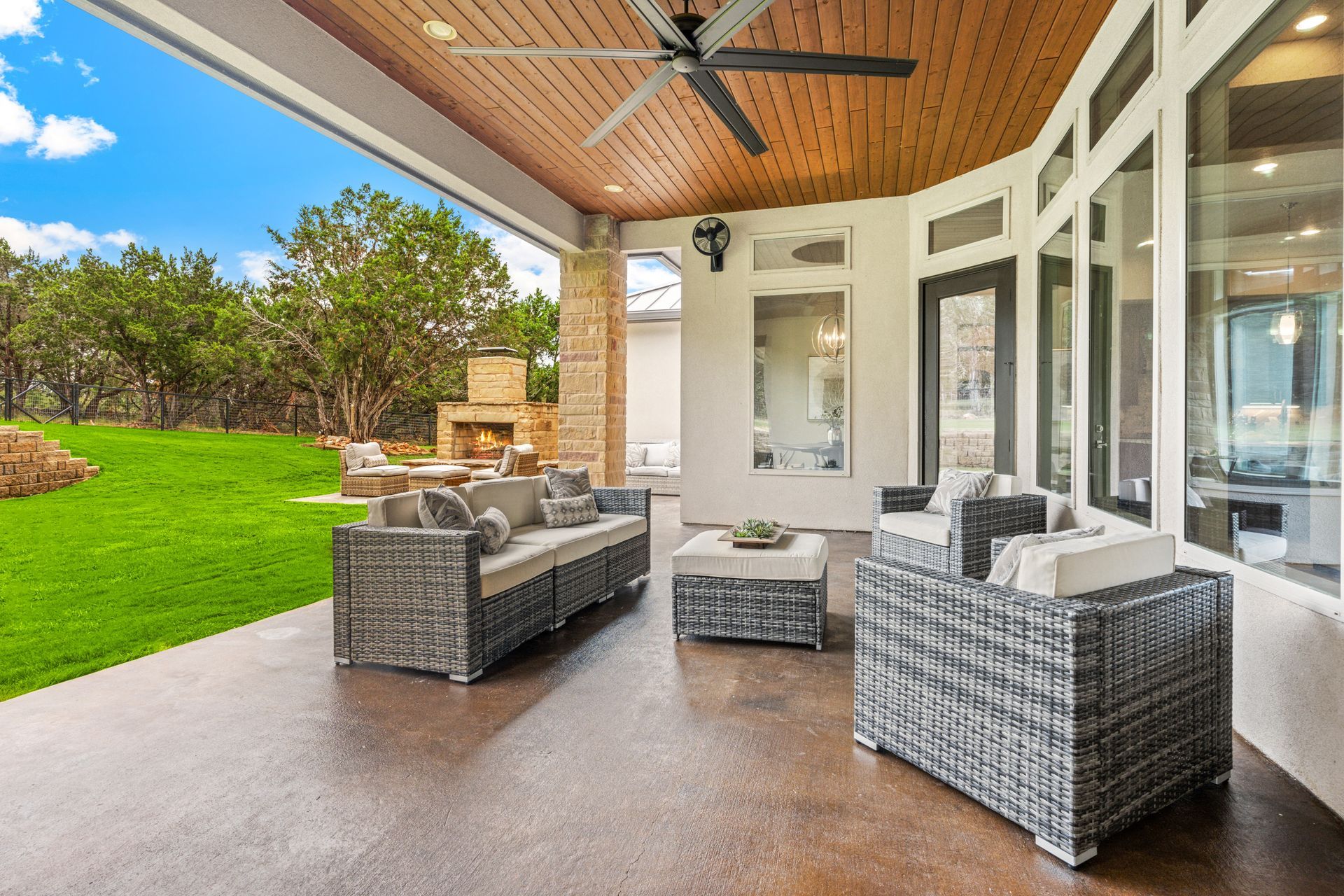 Outdoor patio with wicker furniture, fireplace, and green lawn. Brown ceiling, large windows.