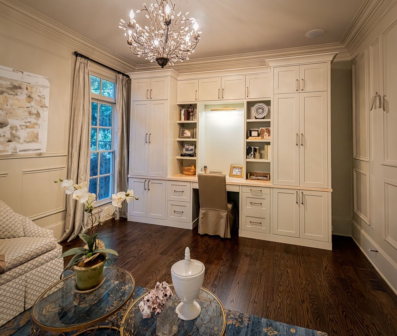 A bright, elegant home office with white cabinetry, a built-in desk, crystal chandelier, and a blue patterned rug.
