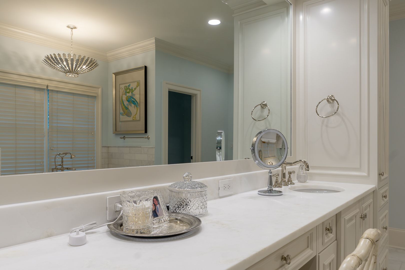 A white bathroom vanity with a long mirror, countertop items, and a round makeup mirror against a light blue wall.