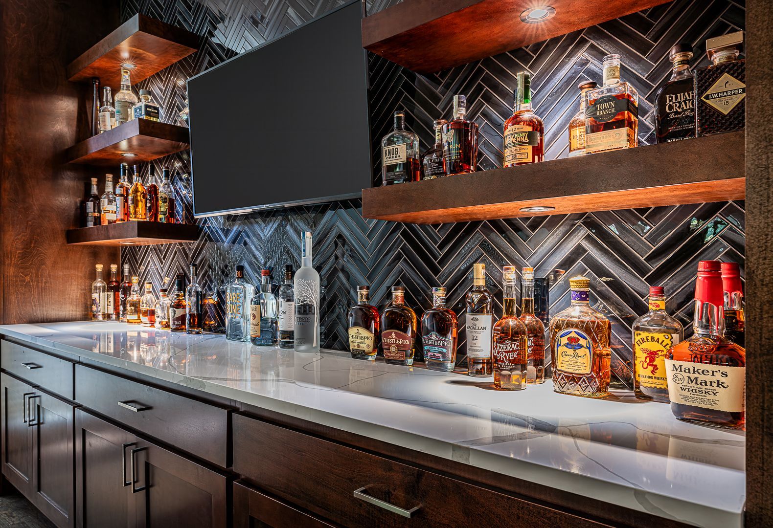 A well-stocked home bar featuring dark wood cabinets, a white countertop, floating wood shelves, and a tiled backsplash.