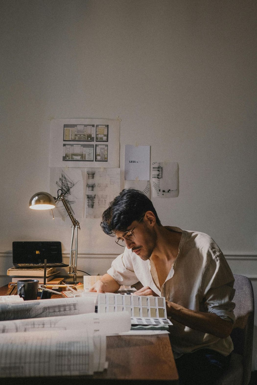 A person in a light shirt focused on architectural drawings at a desk, illuminated by a warm lamp in a dimly lit room.