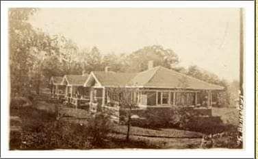 A black and white photo of a house with a porch surrounded by trees.