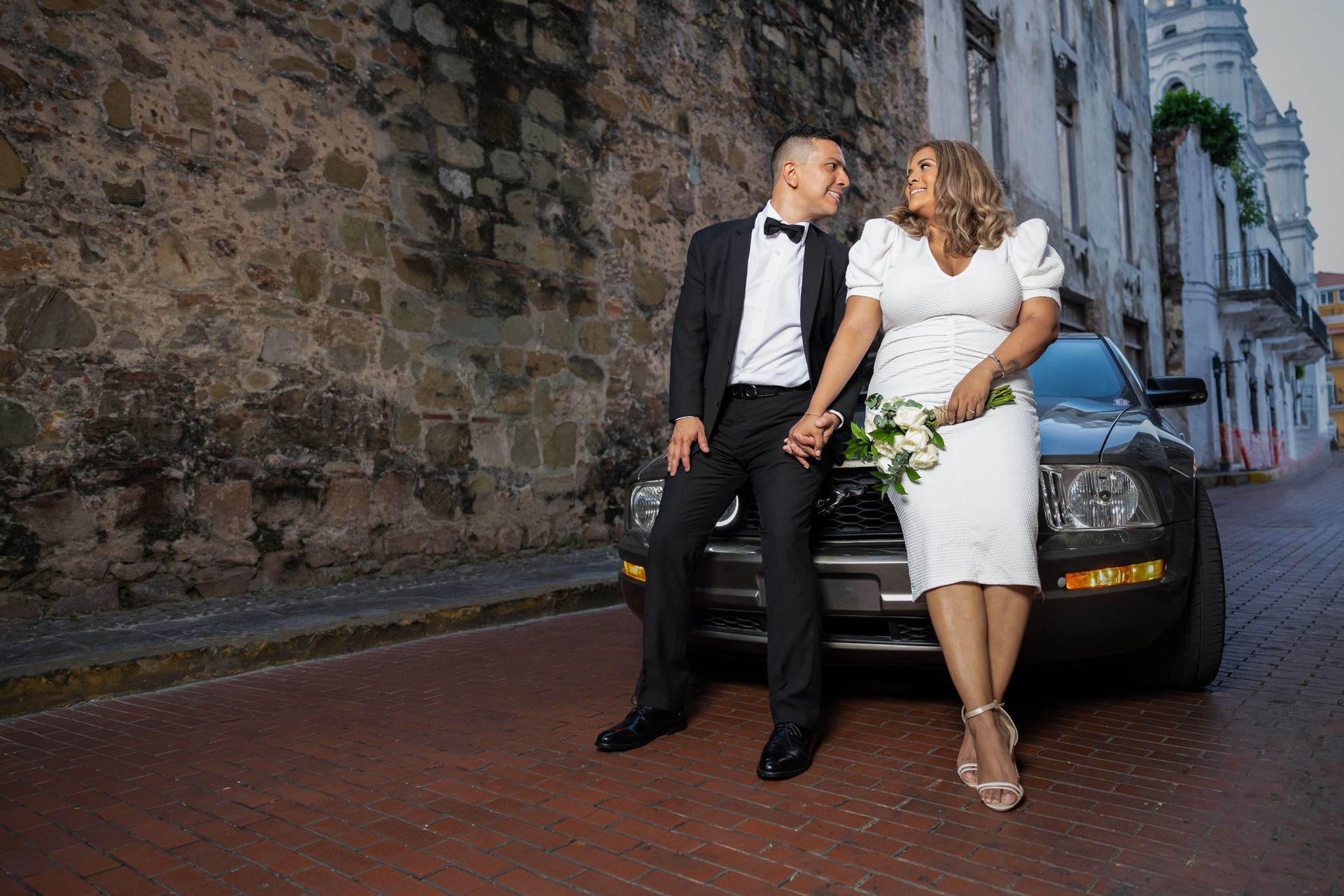 Una novia y un novio están sentados en el capó de un coche tomados de la mano.